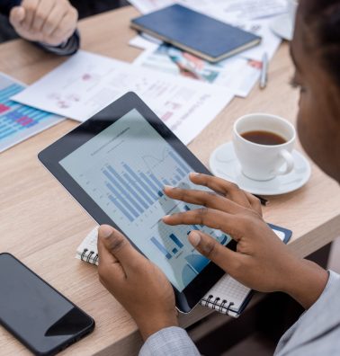 Hand of young African businesswoman scrolling in tablet with charts and diagram on screen while sitting by table