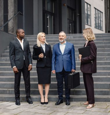 Confident businesspersons standing in front of modern office building. Men and women talking and holding documents. Banking and financial market concept.