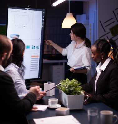 Executive manager woman explaining management statistics working at company strategy overtime in office meeting room. Diverse multi-ethnic businesspeople teamwork brainstorming ideas in evening.