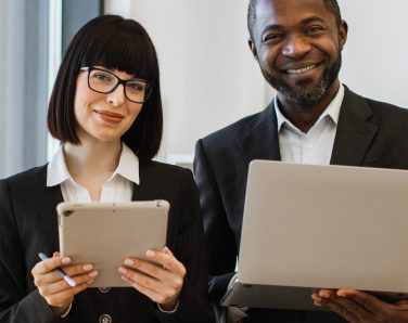 A smiling African American man holds a laptop while a woman with glasses holds a tablet, both dressed in business attire