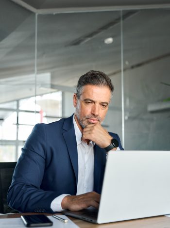 Serious mature business man executive sitting at desk using laptop. Busy professional mid aged businessman ceo financial expert working on computer thinking analyzing investment market risk in office.