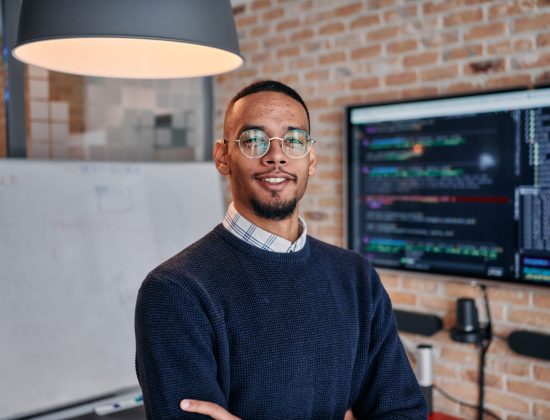 In a modern startup office setting, an African American businessman stands confidently with his colleagues in the background, embodying leadership and teamwork.